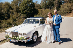 Julia und Stefan in Hochzeitskleidung neben einem weißen geschmückten Oldtimer